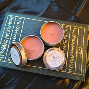 May include: Three round, silver-colored candles with a light brown wax are displayed on a dark green book with gold lettering. One candle has its lid off, revealing the candle's wick. The labels read "Dwarven Library" and "Sweet As Scents Candle Co."