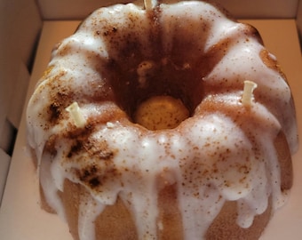 May include: A white frosted bundt cake with cinnamon sprinkles and three wicks. The cake is in a white box.