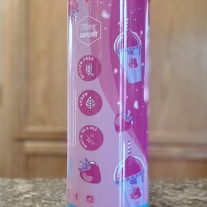 May include: A cylindrical tumbler with a pink and purple strawberry design. The tumbler features the text "200mg Caffeine", and icons indicating it is gluten-free and vegan. The bottom of the tumbler is light blue.