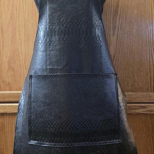 May include: A black apron with a textured, snakeskin-like pattern. The apron has a large front pocket and adjustable straps with metal hardware. The apron is displayed on a black mannequin.