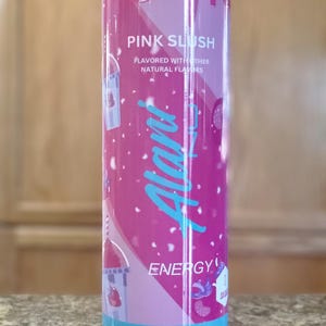 May include: A pink and blue insulated tumbler with the text "PINK SLUSH" and "Alani Energy". The tumbler features a repeating pattern of slushy cups and fruit. The tumbler has a blue base and a clear lid.