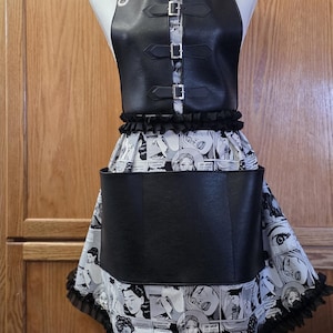 May include: Black apron with a comic book print skirt and a black faux leather bodice. The bodice has three silver buckles and adjustable straps. The skirt features a black and white comic book pattern with a black ruffle trim.