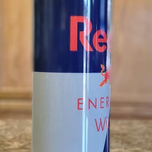 Pu&ograve; includere: Un bicchiere in acciaio inossidabile blu e bianco con un logo Red Bull e il testo "Servito freddo, pu&ograve; darti le ali". Il bicchiere ha anche il testo "Bevanda energetica" e "Prodotto con taurina, vitalizzato con zucchero".