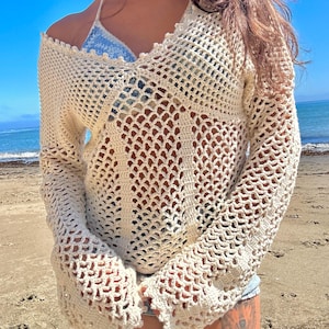May include: A cream-coloured, open-weave crochet sweater with long sleeves. The sweater has a loose fit and a scalloped neckline. The beachwear top is worn over a blue and white bikini top. The background shows a beach and blue sky.