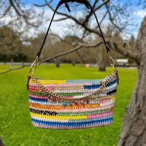 May include: A multi-coloured crocheted handbag with a black strap and gold-tone chain detail. The bag features a variety of colours, including pink, yellow, blue, and black. The chain is adorned with small charms. The bag is suspended from a tree branch.