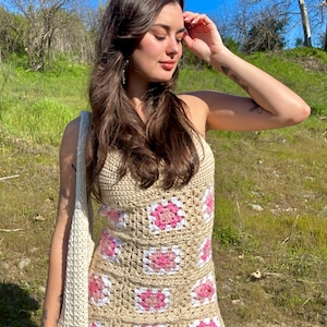 May include: A woman wearing a crochet dress with pink and white squares. The dress is sleeveless and has a fitted silhouette. The woman is standing in a grassy field with a blue sky in the background.