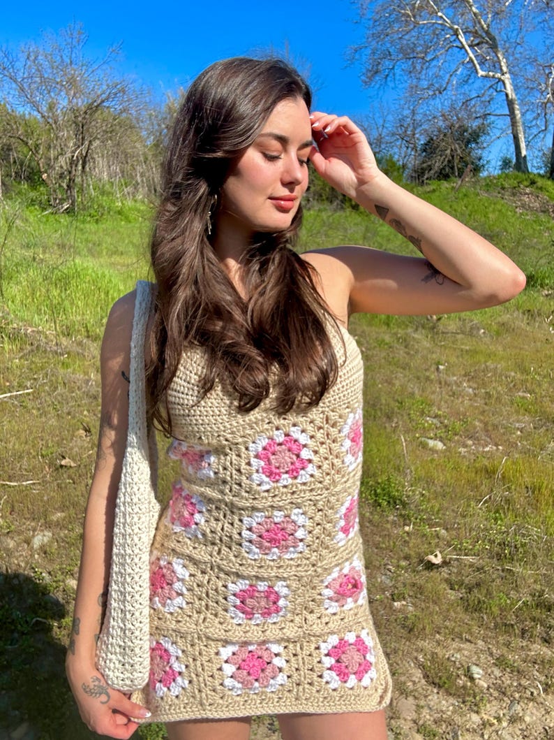 May include: A woman wearing a crochet dress with pink and white squares. The dress is sleeveless and has a fitted silhouette. The woman is standing in a grassy field with a blue sky in the background.