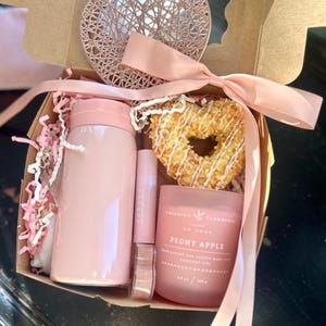 May include: A gift box with a pink theme, containing a pink tumbler, lip gloss, a candle, and a heart-shaped pastry. The box is tied with a pink ribbon and includes a decorative heart.