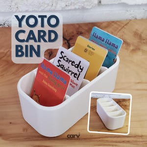 May include: A white YOTO CARD BIN with four compartments, filled with children's books. The books feature titles such as "Llama Llama" and "Scaredy Squirrel." The bin is on a wooden surface, with a smaller image of the bin in the corner.