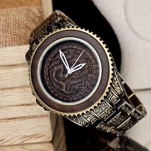 May include: A wooden wristwatch with a dark brown face featuring a carved dragon design. The watch has gold-colored metal accents and a matching band. The watch is displayed on a reflective surface.
