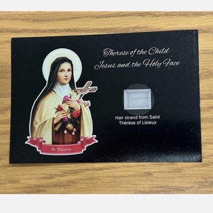 May include: A black card with an image of St. Thérèse, holding flowers and a crucifix. The card has the text "Therese of the Child Jesus and the Holy Face" and "Hair strand from Saint Thérèse of Lisieux."