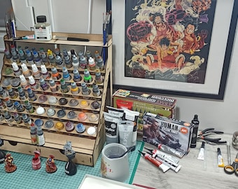 May include: A wooden paint rack with multiple rows of small paint bottles, a variety of acrylic paints, and miniature figures on a green surface.