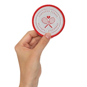 Puede incluir: Un parche redondo con borde rojo y centro blanco. El parche presenta el texto "TORTURED TENNIS PLAYERS DEPARTMENT" alrededor del borde. En el centro, dos raquetas de tenis cruzadas y un símbolo de corazón roto.