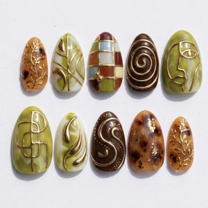 May include: A set of ten almond-shaped press-on nails featuring various designs including gold leaf, metallic gold line art, abstract swirls, geometric grids, and a face silhouette in shades of olive green, brown, amber, and cream.