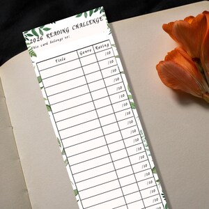 May include: A white bookmark with a green leaf border and the text "2026 READING CHALLENGE." The bookmark has spaces to record book titles, genres, and ratings. A flower with orange petals is in the background.