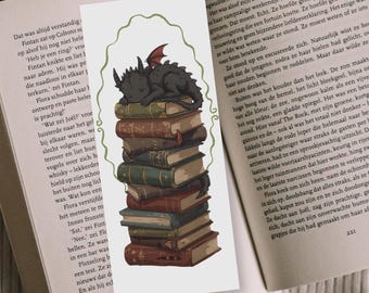 May include: Bookmark featuring a black dragon with red wings sleeping atop a stack of old books. The bookmark has a green border and is set against the backdrop of an open book.