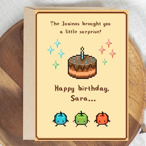 May include: A rectangular greeting card with a light yellow background featuring pixel art of a chocolate cake with a single candle, surrounded by colorful sparkles. The text reads: The Junimos brought you a little surprise! Happy birthday, Sara... with three small pixelated apple-like creatures at the bottom.