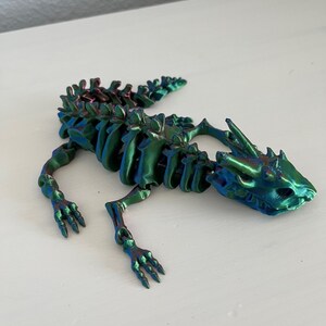 May include: A 3D printed skeleton of a lizard with a green, blue, and purple iridescent finish. The skeleton is posed in a walking position with its head turned to the side.