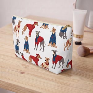 May include: A white rectangular zippered pouch featuring a repeating pattern of grey and tan greyhounds wearing various red and blue sweaters, scarves, and hats. The pouch has a gold-toned metal zipper closure along the top edge.