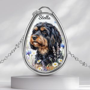 May include: Silver-toned pendant necklace featuring a watercolor portrait of a dog with black and tan fur, surrounded by flowers. The name "Stella" is written above the dog. The pendant hangs from a delicate chain.