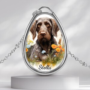 Könnte beinhalten: Eine silberfarbene Halskette mit Anhänger, die ein Aquarellporträt eines braunen und grauen Hundes namens Stella zeigt, umgeben von bunten Wildblumen. Der tropfenförmige Anhänger hängt an einer filigranen Kette.