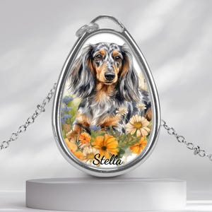 May include: Silver pendant necklace with a watercolour illustration of a long-haired dachshund. The dog is set against a backdrop of yellow, orange, and blue flowers. The name "Stella" is written at the bottom of the pendant.