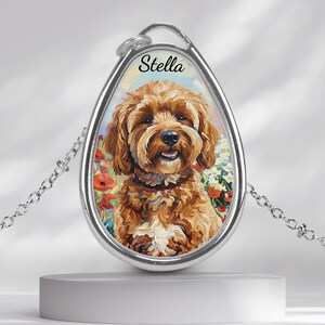 Puede incluir: Collar con colgante plateado con un retrato pintado de un perro marrón, con el nombre "Stella" encima. El perro está sobre un fondo floral. El colgante cuelga de una delicada cadena.