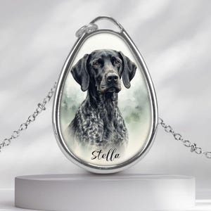 Op de afbeelding: Een zilverkleurige ovale hanger aan een ketting met een aquarelillustratie van een zwart-witte hond met vlekken. De naam Stella staat in zwarte cursieve letters onderaan het portret, dat is gevat in een metalen omlijsting.