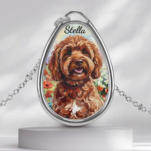 May include: Silver pendant necklace with a portrait of a brown dog, named "Stella". The dog is set against a floral background. The pendant hangs from a delicate chain. The pendant is approximately 3 cm in length.