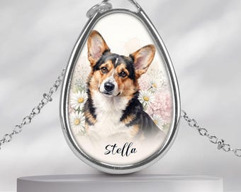 May include: A silver-toned oval pendant necklace featuring a watercolour-style illustration of a black, tan, and white Corgi dog surrounded by white daisies and pink flowers, with the name Stella printed in black script lettering below the dog.