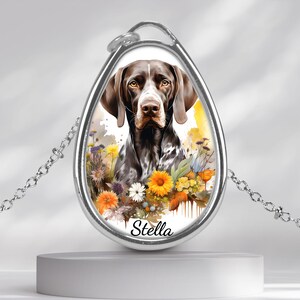 May include: Silver-toned pendant necklace featuring a watercolor-style portrait of a brown and white dog surrounded by colorful flowers. The dog's name, "Stella," is printed at the bottom of the pendant.
