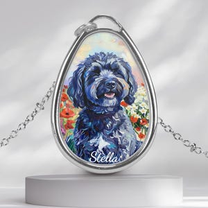 Può includere: Collana con pendente argentato raffigurante un ritratto dipinto di un cane nero con un'espressione felice, circondato da fiori. Il pendente a forma di goccia ha il nome "Stella" in basso. La catena è delicata e argentata.