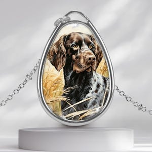 Può includere: Una collana con pendente color argento con un ritratto dipinto di un cane marrone e bianco. Il cane è ambientato su uno sfondo di erba alta e dorata. Il pendente è a forma di goccia e pende da una delicata catena.