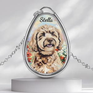 May include: Silver-toned pendant necklace featuring a smiling dog portrait with the name "Stella" above. The dog is light brown with a fluffy coat, set against a floral background. The pendant hangs from a delicate chain.