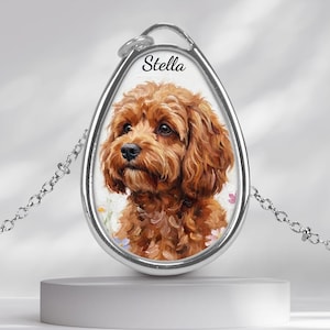 Puede incluir: Un collar con colgante plateado con un retrato pintado de un perro marrón, con el nombre "Stella" encima. El perro está sobre un fondo blanco con detalles florales. El colgante cuelga de una delicada cadena. Un accesorio con temática de mascotas.