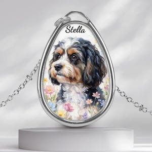 May include: A silver-toned pendant necklace featuring a painted portrait of a dog with black, brown, and white fur, surrounded by colorful flowers. The name "Stella" is written above the dog's head. The pendant hangs from a delicate chain.
