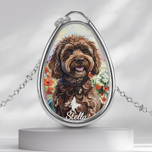 Könnte beinhalten: Eine silberfarbene Halskette mit Anhänger, die ein gemaltes Porträt eines braunen Hundes mit fröhlichem Gesicht zeigt. Der Hund ist vor einem Hintergrund aus Blumen platziert. Der Name "Stella" steht unten auf dem Anhänger.
