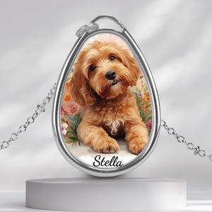 May include: A silver pendant necklace with a watercolour portrait of a light brown, fluffy dog. The dog is set against a floral background, with the name "Stella" at the bottom. The pendant is a teardrop shape and is suspended on a silver chain.