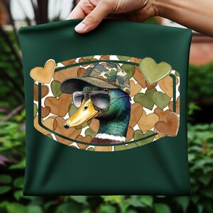 May include: A dark green cushion cover featuring a cartoon duck wearing sunglasses and a camouflage hat. The duck is inside a brown oval frame with heart-shaped accents in shades of brown and green. The cushion cover is held by a person.