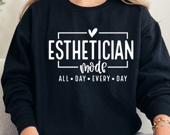 Op de afbeelding: Zwart sweatshirt met witte tekst die "Esthetician mode all day every day" luidt met een klein hartje boven het woord "Esthetician".