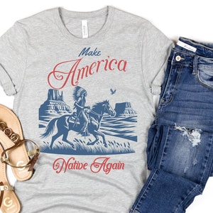 May include: Gray t-shirt with a blue and red graphic of a Native American riding a horse in a desert landscape. The text "Make America Native Again" is printed below the graphic.