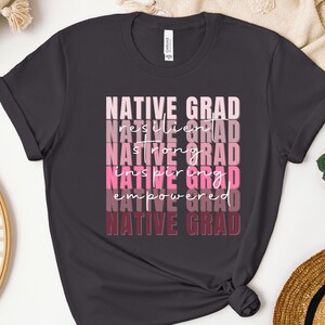 May include: A dark gray t-shirt with the text "NATIVE GRAD" repeated in a gradient of pink and burgundy colors. The text is stacked vertically with each word slightly offset from the one above it.