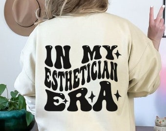 Op de afbeelding: Een crèmekleurig sweatshirt met de tekst "IN MY ESTHETICIAN ERA" in zwart gedrukt op de achterkant. De tekst is in een retro-lettertype met sterren.