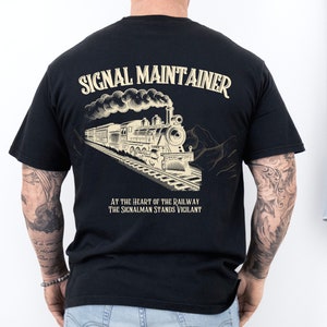 Puede incluir: Camiseta negra con una ilustración vintage de un tren y el texto "SIGNAL MAINTAINER" en amarillo. El texto "AT THE HEART OF THE RAILWAY THE SIGNALMAN STANDS VIGILANT" está impreso debajo de la ilustración.