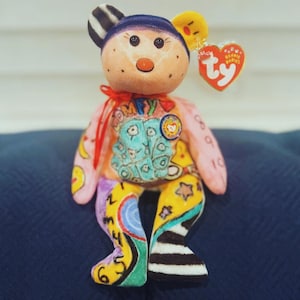 Puede incluir: Un colorido peluche de oso Ty Beanie Baby con una etiqueta en forma de corazón. El oso tiene una cara marrón claro, un gorro azul y extremidades multicolores con varios diseños, incluyendo números, estrellas y rayas. El pecho presenta la palabra "COMFY".
