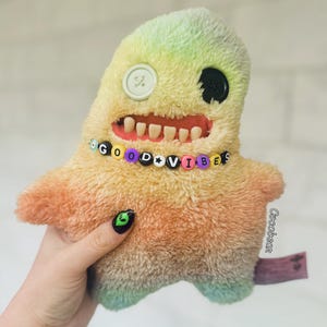 Op de afbeelding: Een pluche speelgoed in de vorm van een monster met een regenboogkleurige gradiënt. Het speelgoed heeft een grote, tandenrijke grijns, één knoopoog en een kralenketting met de tekst "GOOD VIBES".