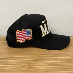 May include: A black baseball cap with a white embroidered American flag on the left side and the text "45**" and "MAKE" in gold embroidery on the right side.