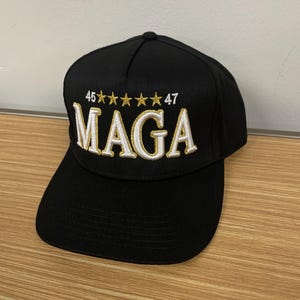 May include: A black baseball cap with a white embroidered "MAGA" logo and gold stars. The numbers "46" and "47" are also embroidered in gold.