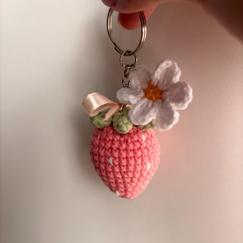 May include: A crochet strawberry keychain featuring a pink textured fruit body with small white embroidered accents, a light green top, a small beige ribbon bow, and a white flower with a yellow center attached to a silver metal keyring.