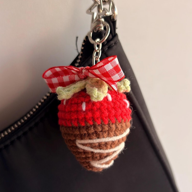 May include: A crocheted strawberry keychain featuring a red upper section with white embroidered seeds, a brown lower section with white yarn drizzle, a light green crocheted leaf top, and a red and white gingham ribbon bow attached to a metal ring.